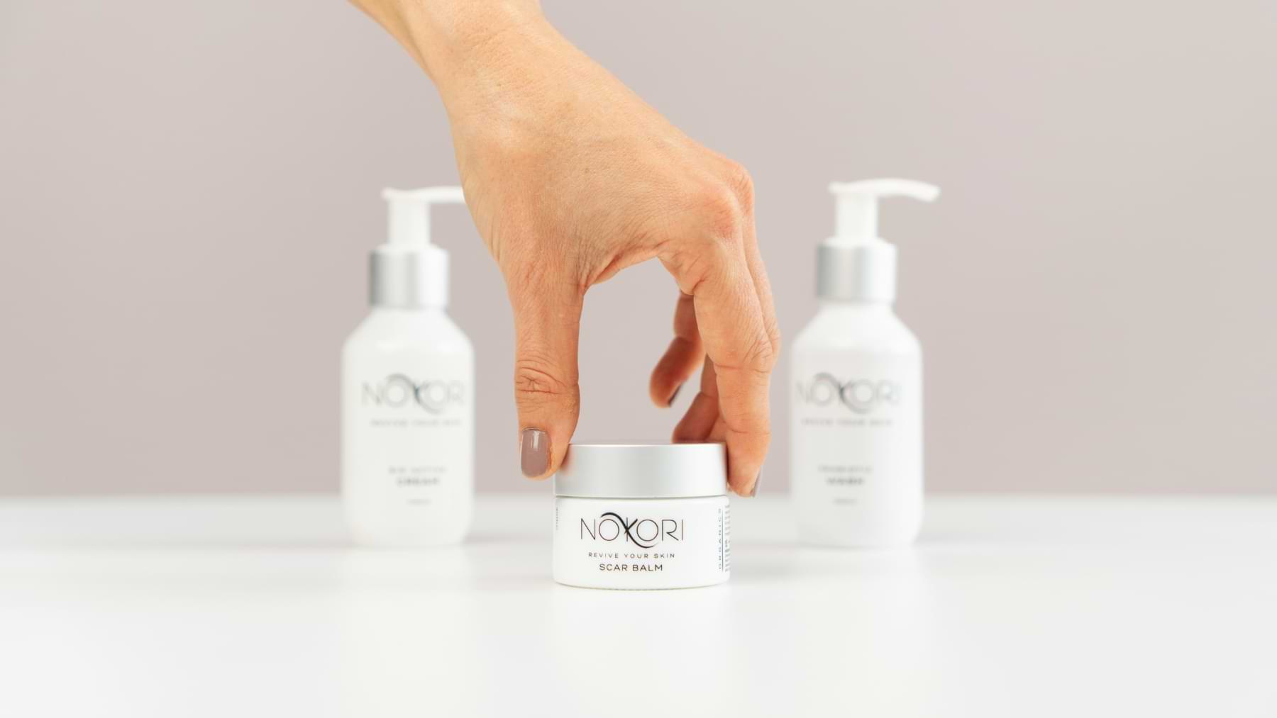 A hand reaching to pick up a Nokori Scar Balm jar, with two Nokori skincare pump bottles blurred in the background.