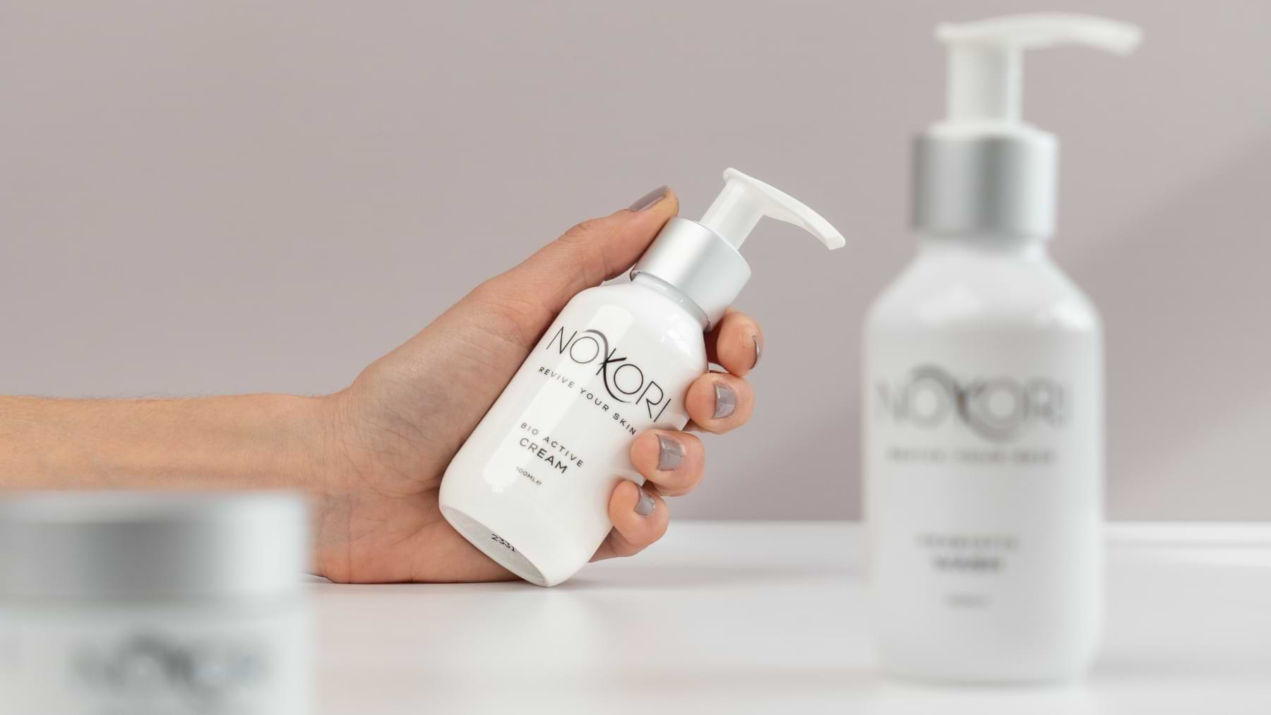 A hand grasping the Nokori Bio Active Cream bottle, with additional product bottles blurred in the background.