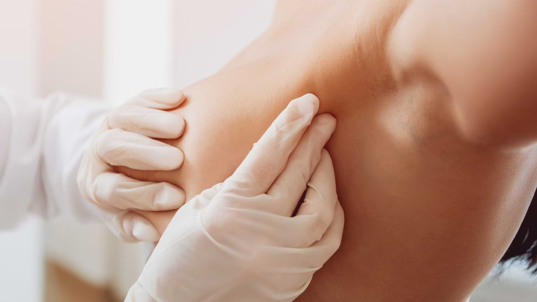 A close-up of gloved hands gently examining the side of a woman's breast during a medical check-up.