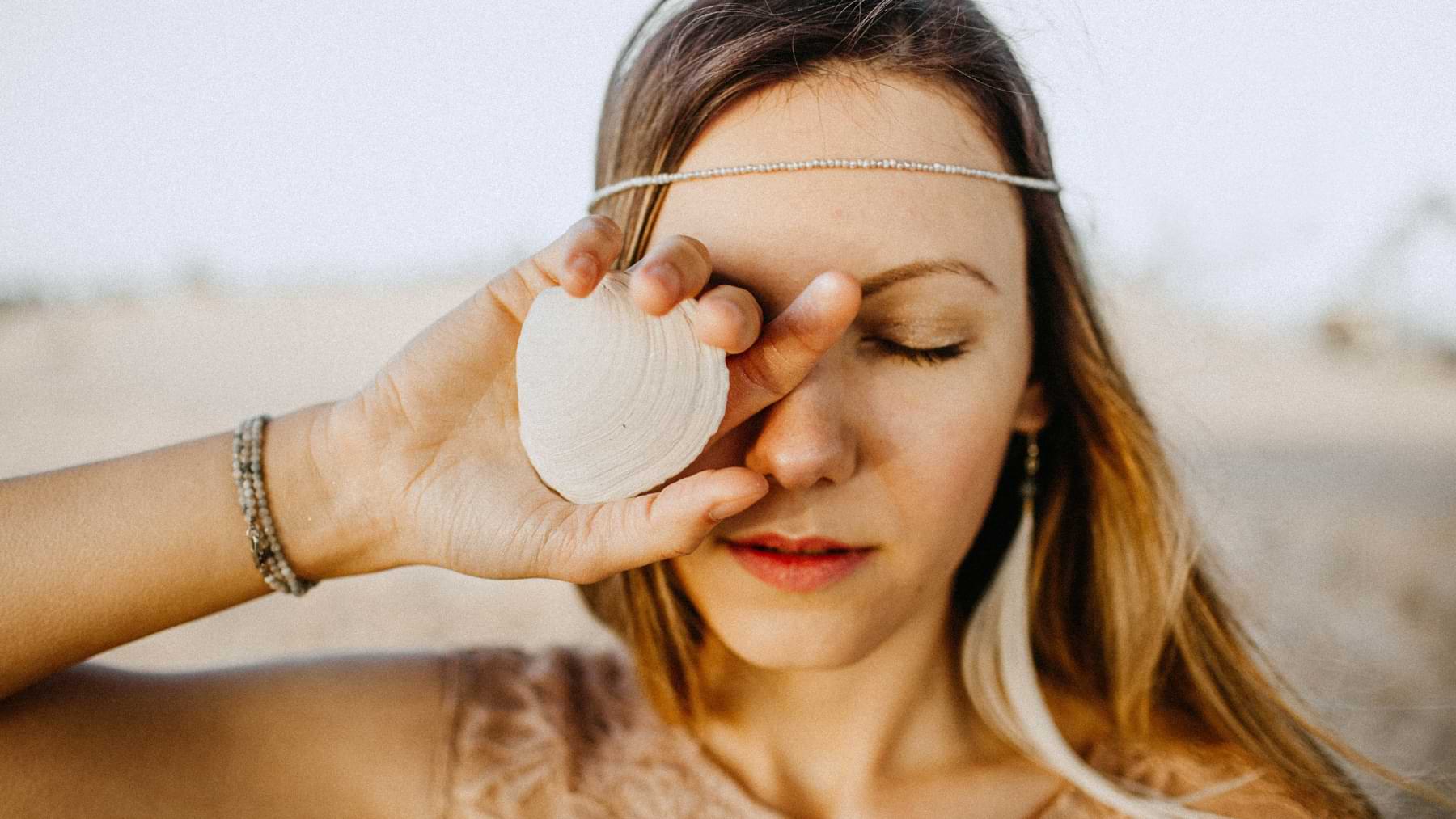 Woman standing outdoors with closed eyes, holding a white seashell in front of one eye.