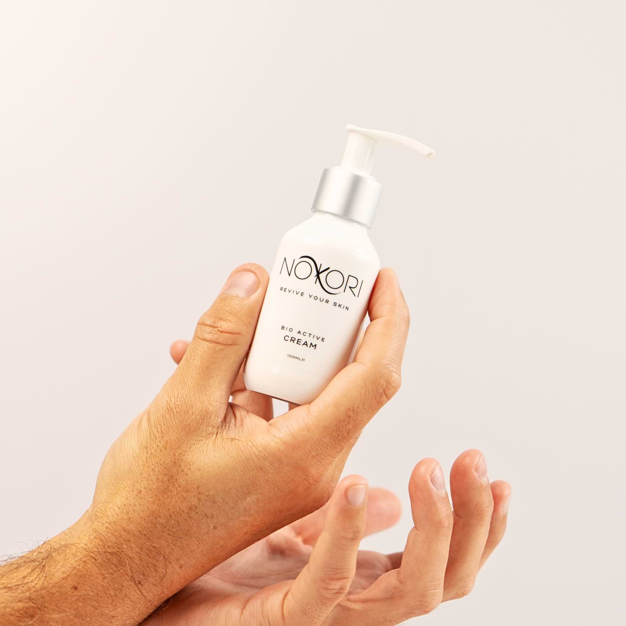 Hands holding a bottle of Nokori Bio Active Cream against a clean, neutral background