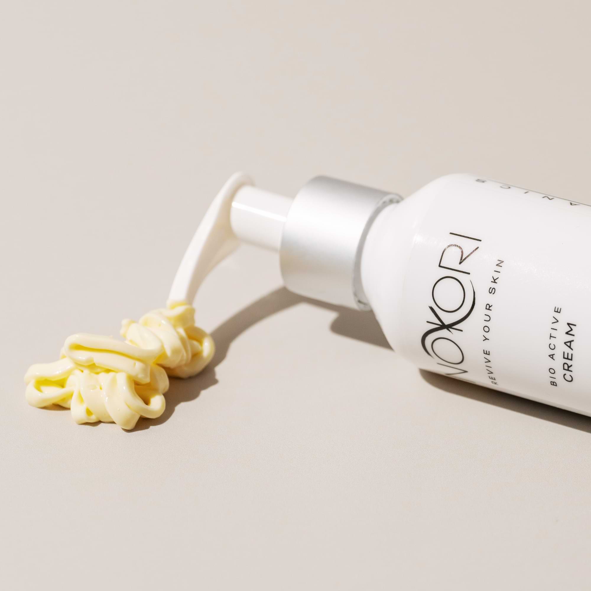 Nokori Bio Active Cream bottle laying on a neutral surface with a generous amount of cream swirled beside it.