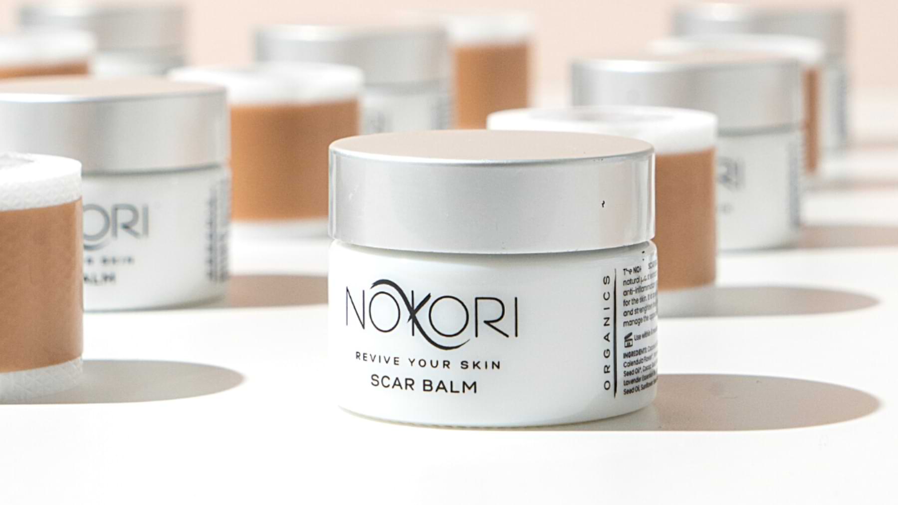 Multiple jars of Nokori Scar Balm and rolls of silicone tape displayed on a white surface with soft shadows.