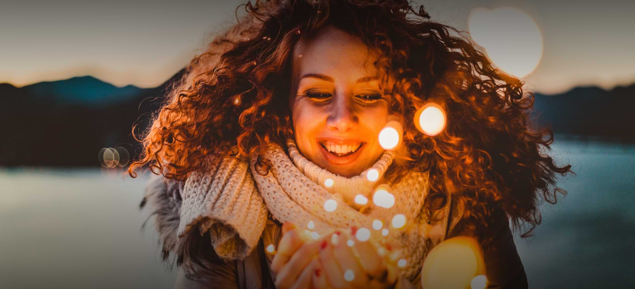 A woman with curly red hair smiles warmly while holding glowing string lights in her hands, with a lake and mountains blurred in the background at dusk.