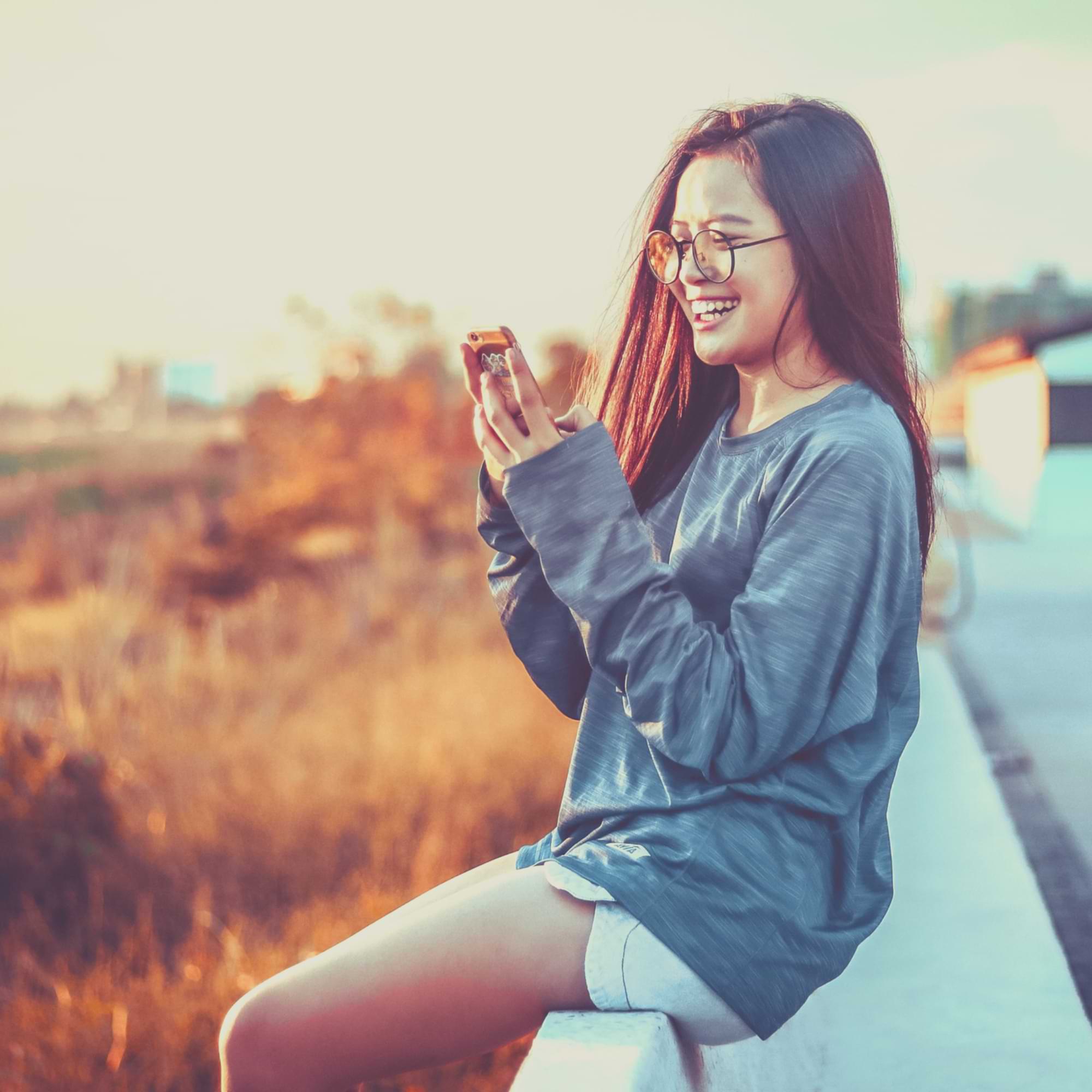 Young woman smiling at her phone outdoors during golden hour, enjoying a moment of connection.