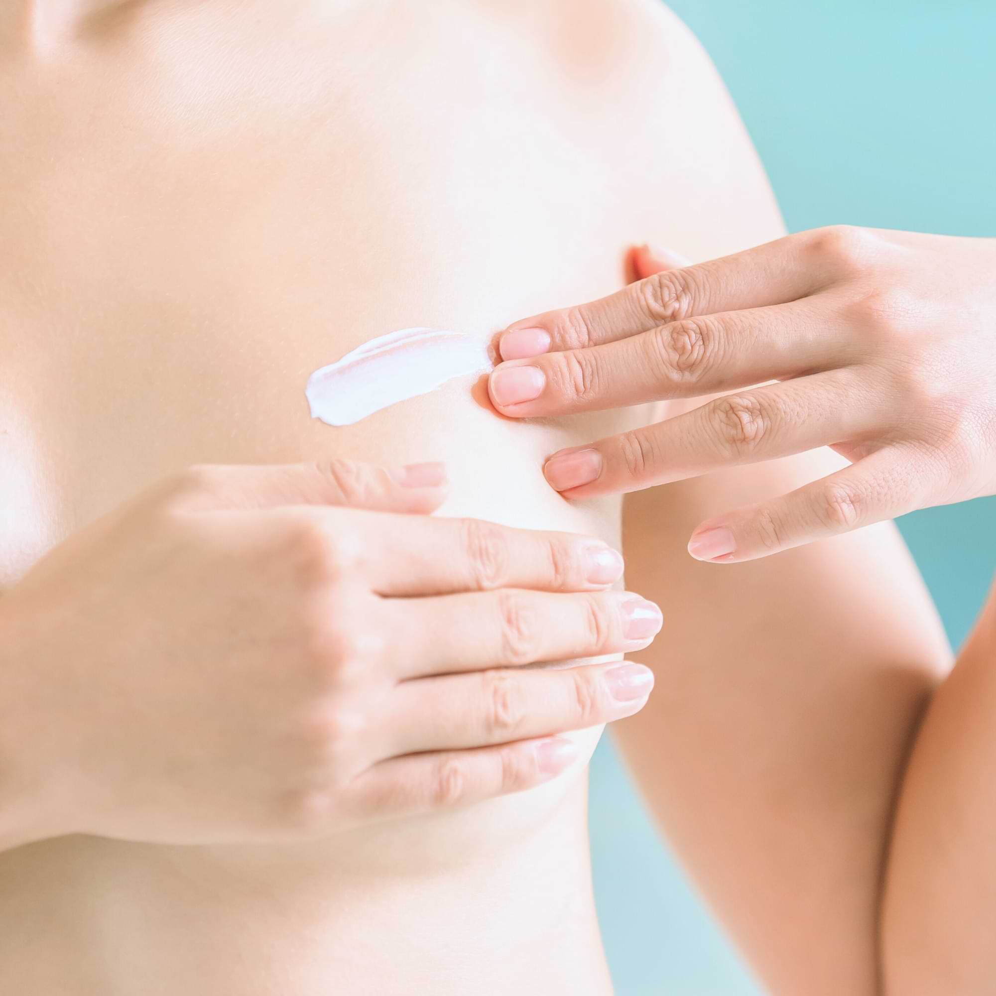 Close-up of a woman applying a streak of cream to the side of her breast with gentle fingertips, against a soft blue background.