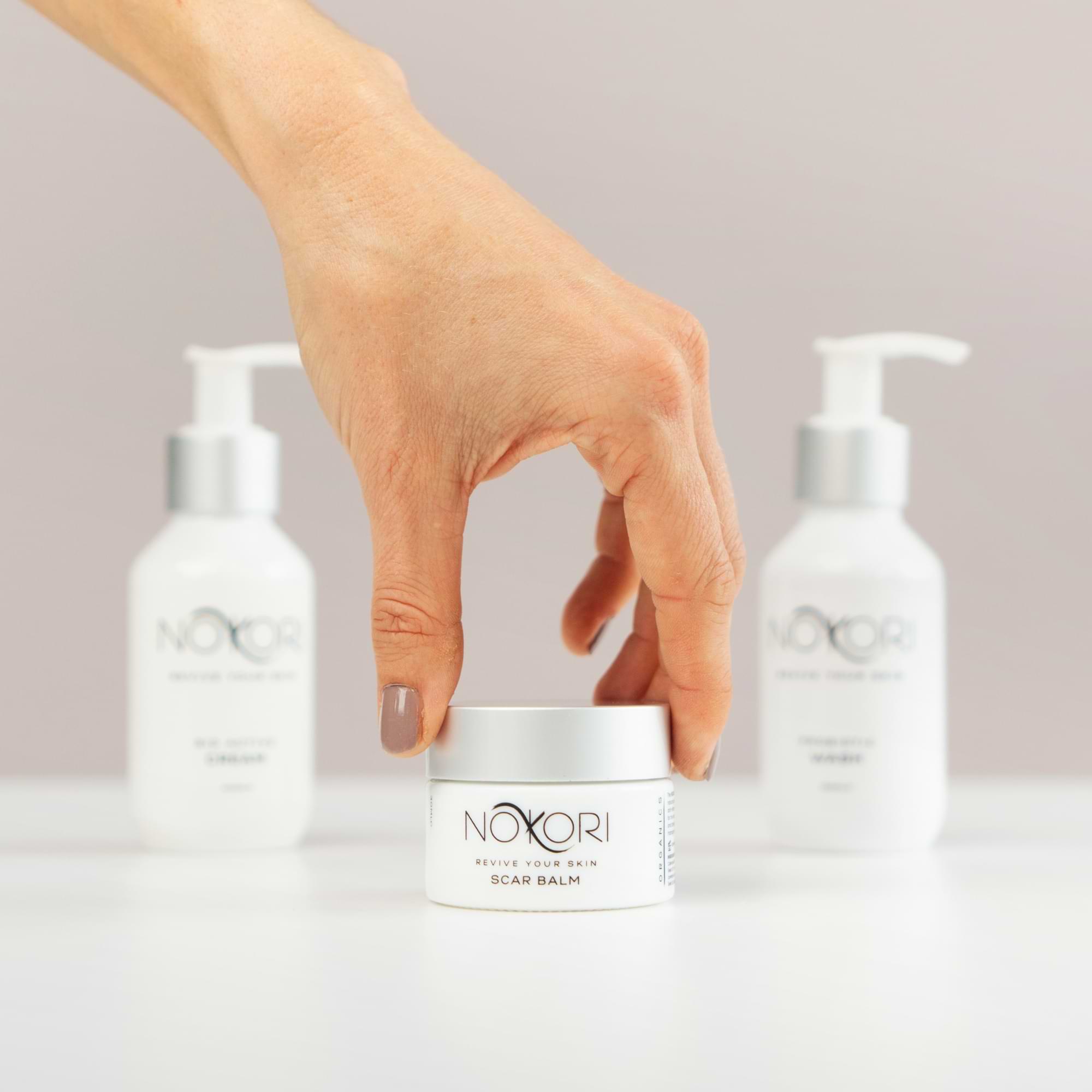 Hand reaching for Nokori Scar Balm, with the Bio Active Cream and Probiotic Wash bottles blurred in the background.