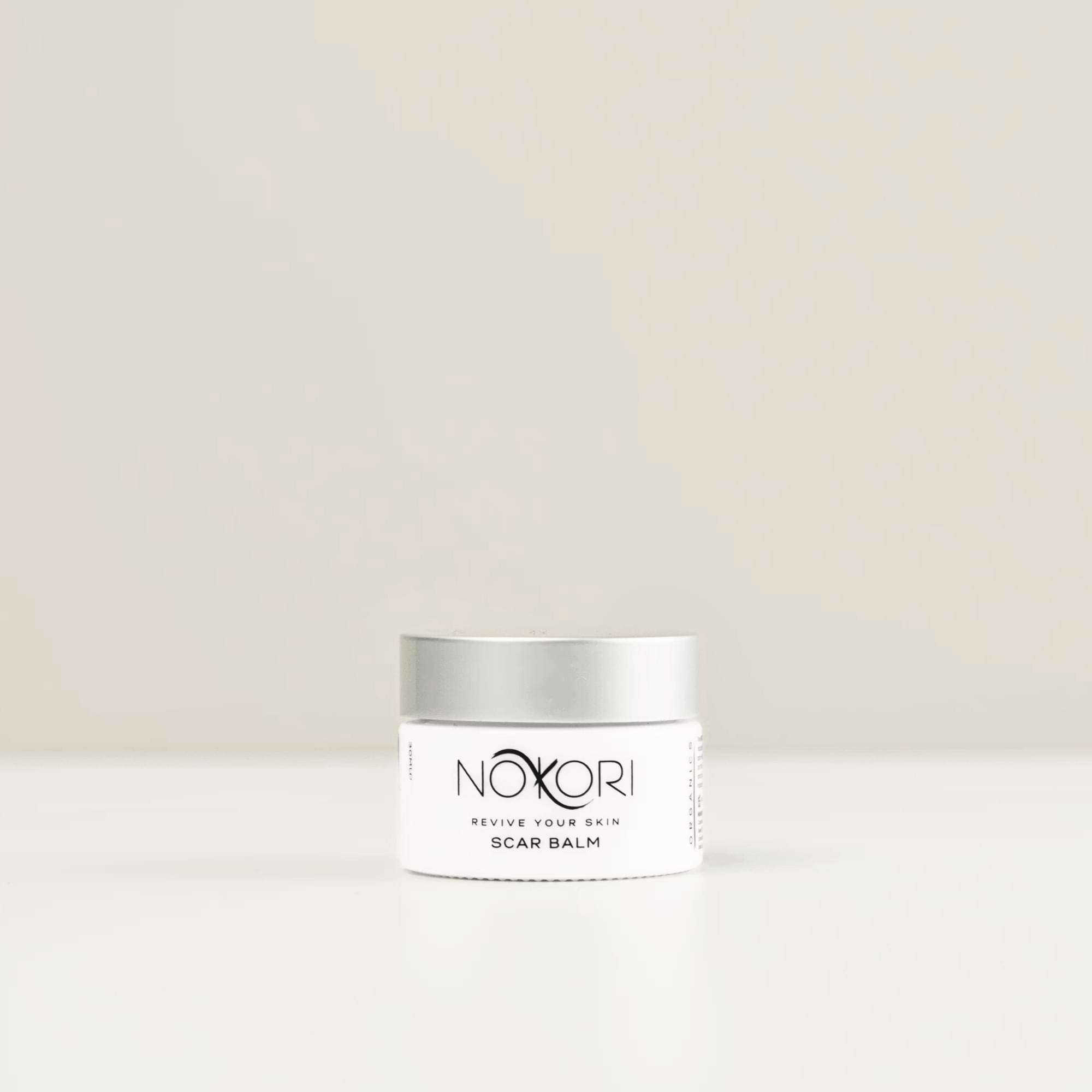 A white jar of Nokori Scar Balm with a silver lid is placed against a soft beige background.