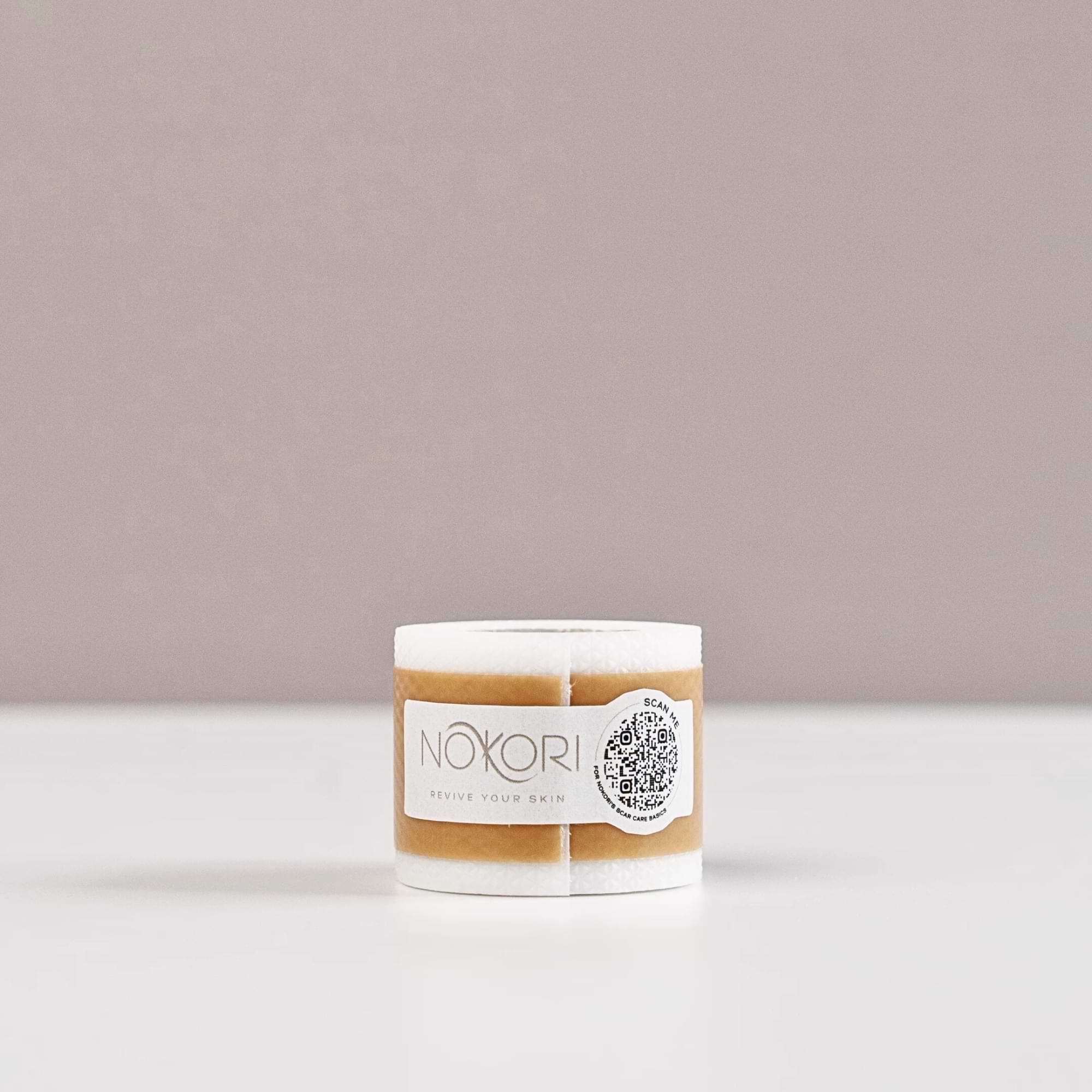 A single roll of Nokori Scar Tape, featuring a tan silicone layer and a QR code on the label, sits against a warm grey background.
