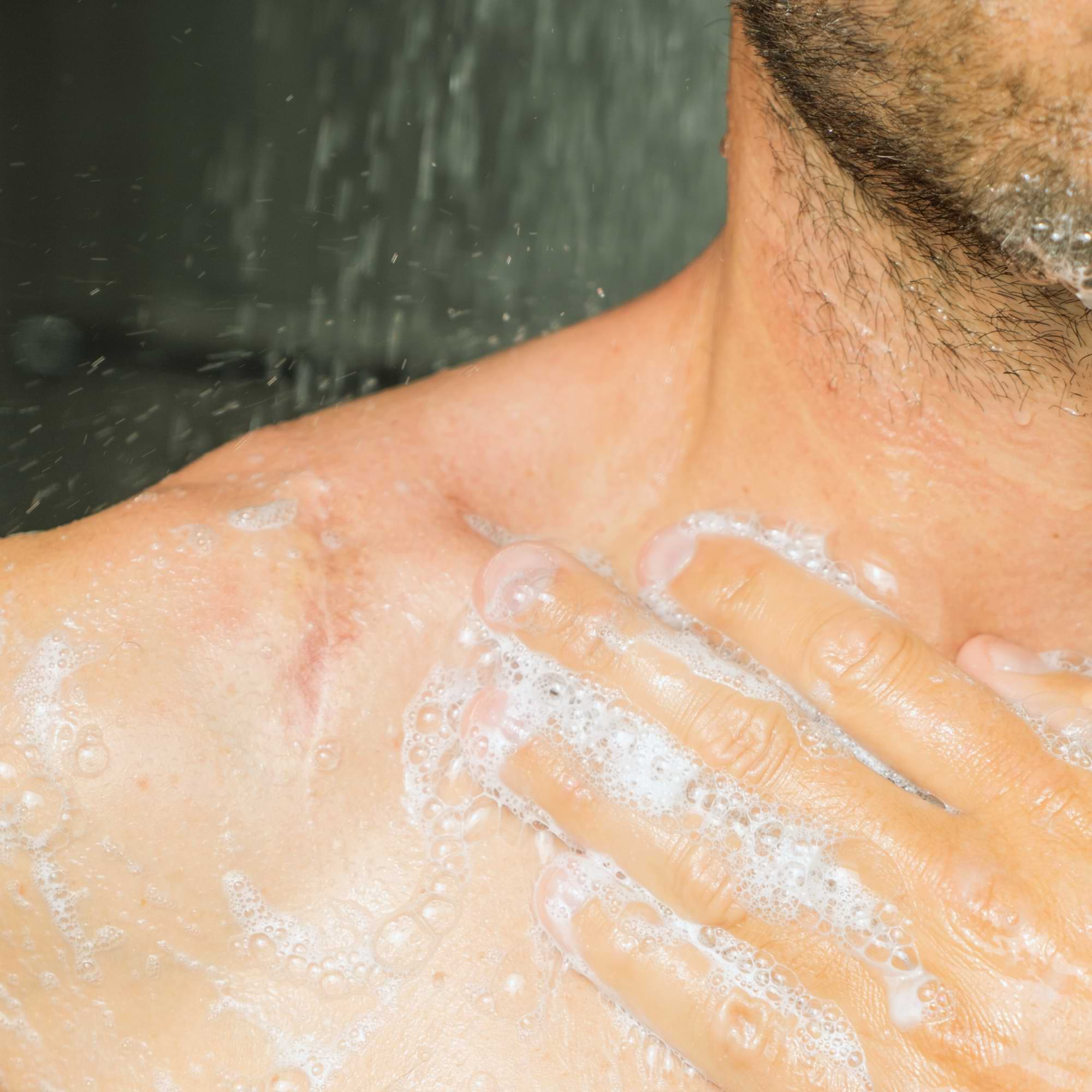 Man washing his upper chest and shoulder in the shower, with visible soap lather and healed scar.