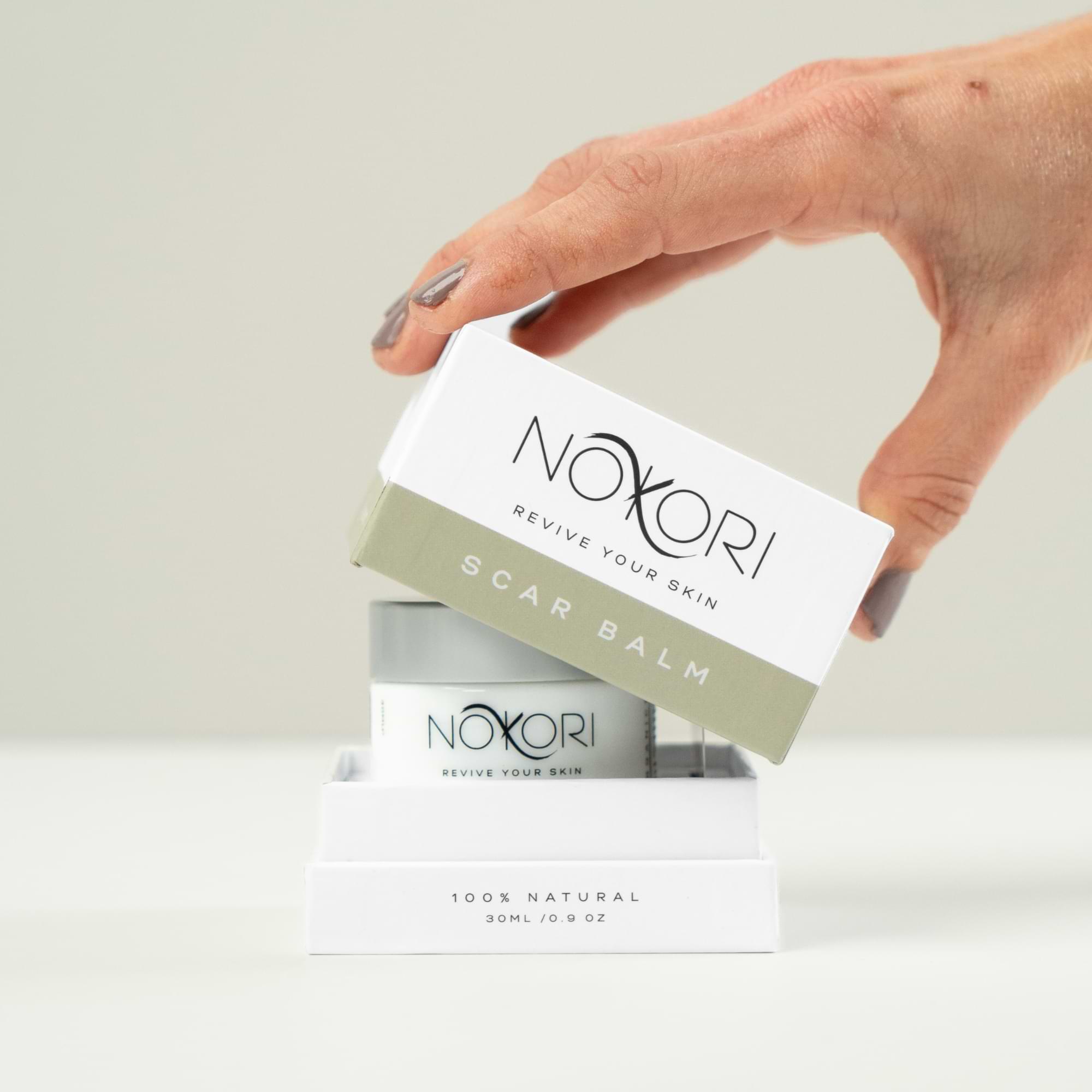 Hand removing the lid of a Nokori Scar Balm box, revealing the jar inside.